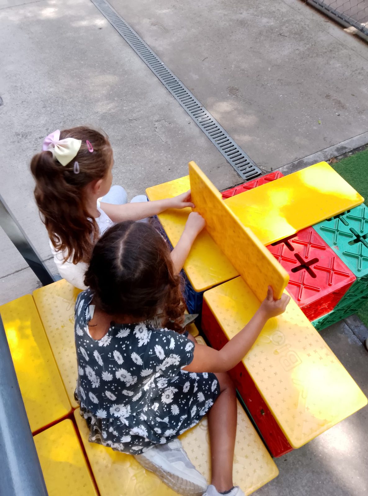 Dues nenes aixequen i encaixen peces grogues i vermelles de gran format sobre un banc del pati.