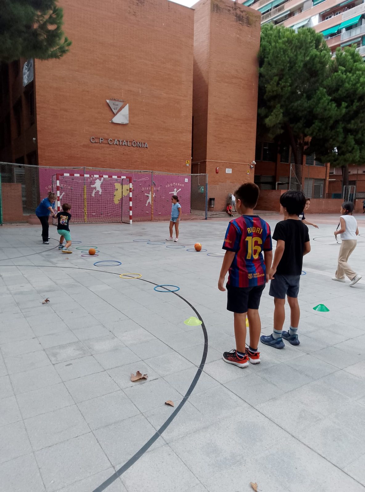 Infants jugant amb pilotes i cèrcols a la pista multiesportiva.