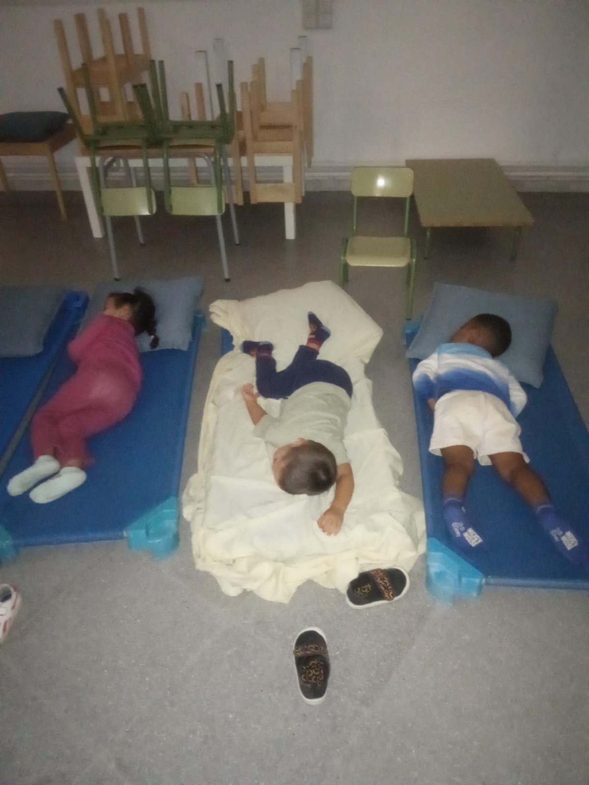Tres infants descansen sobre màrfegues al terra d’una aula amb les cadires apilades.