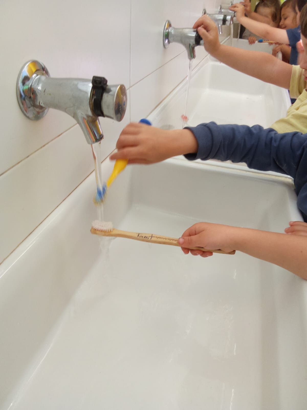 Diverses mans infantils posen pasta al raspall en una pica llarga del lavabo.