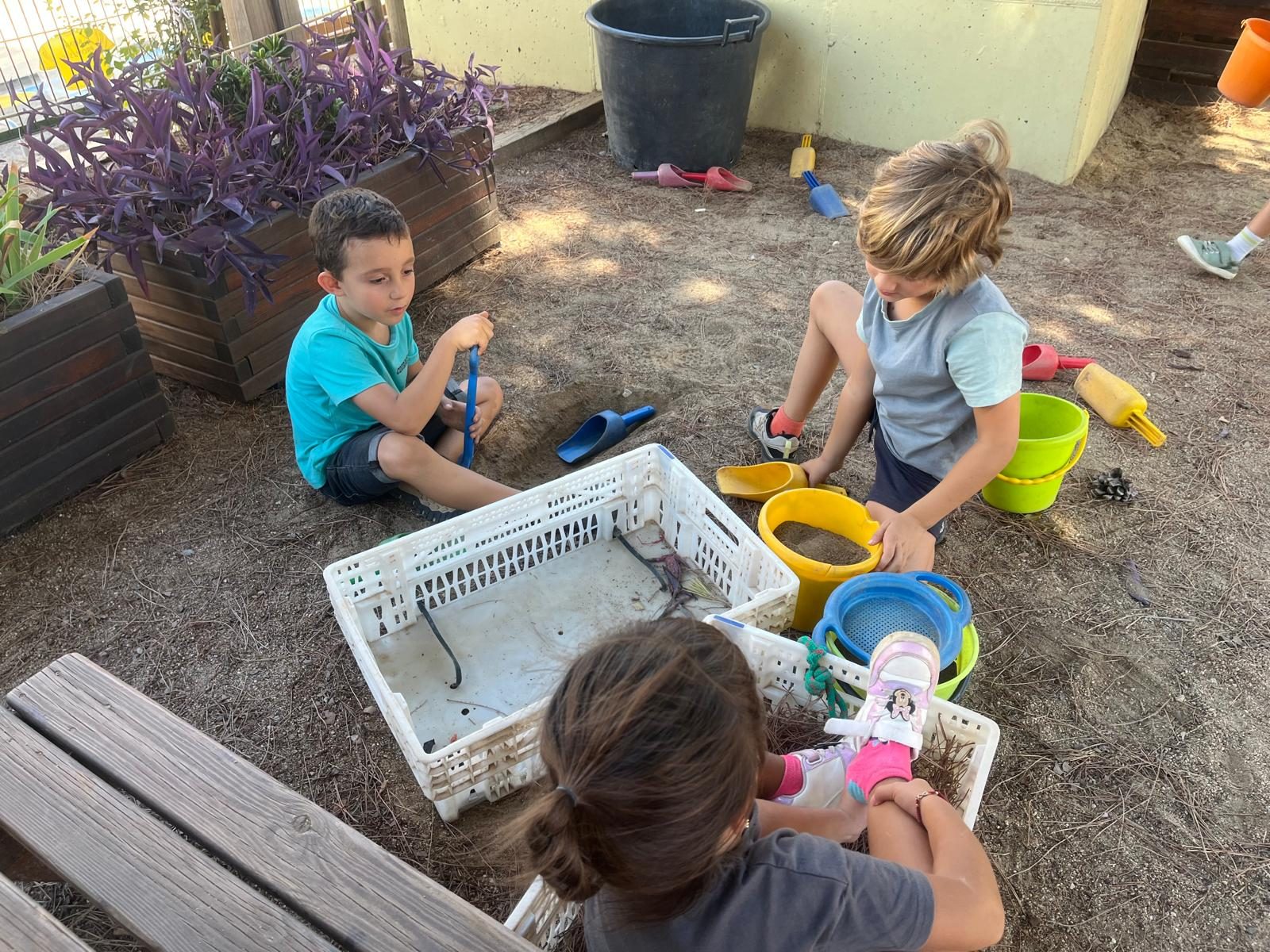 Infants asseguts a terra remenen sorra amb galledes i caixes de plàstic.