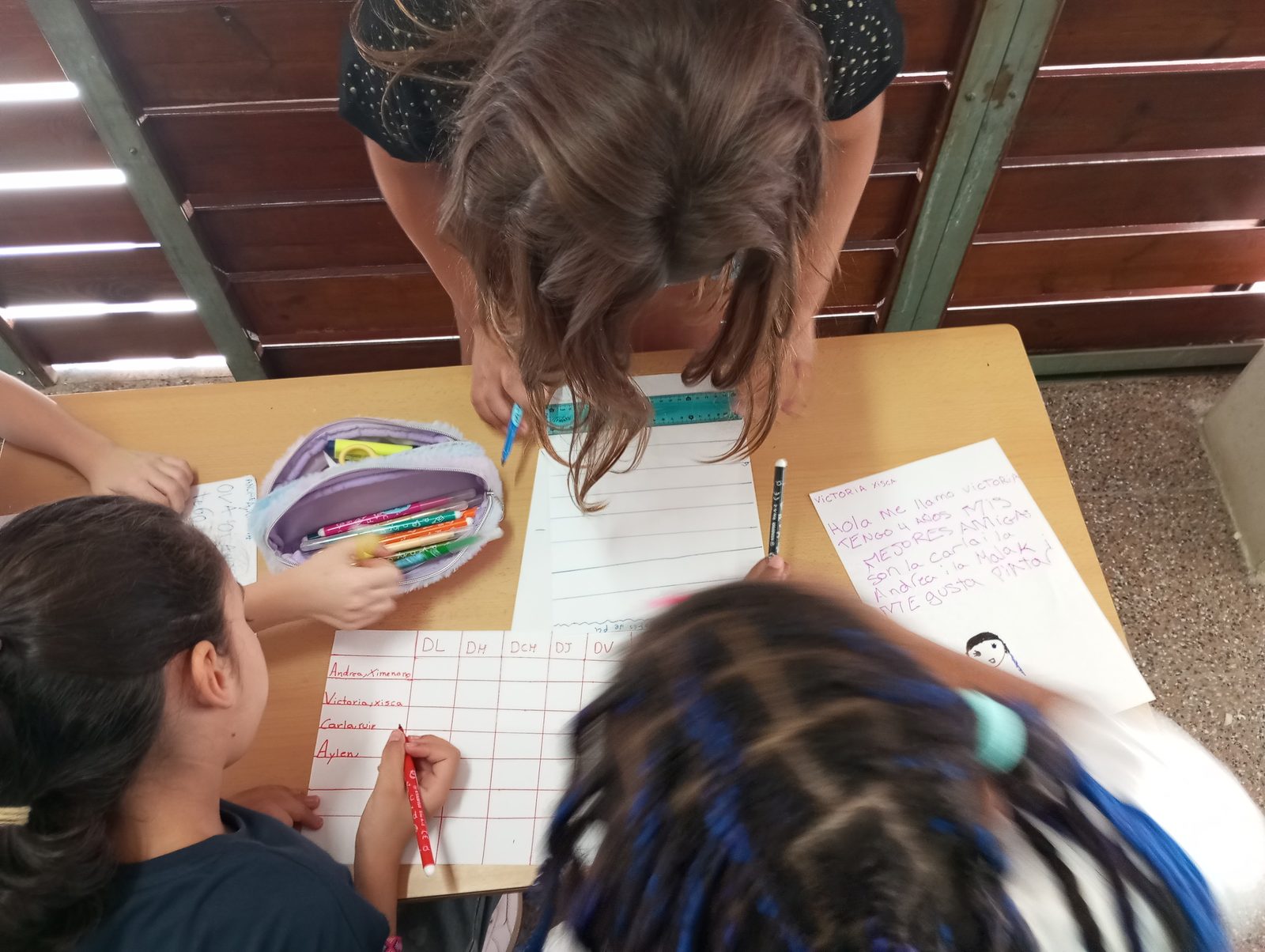 Una educadora i diversos infants escriuen amb retoladors de colors un full amb els dies de la setmana en columnes.