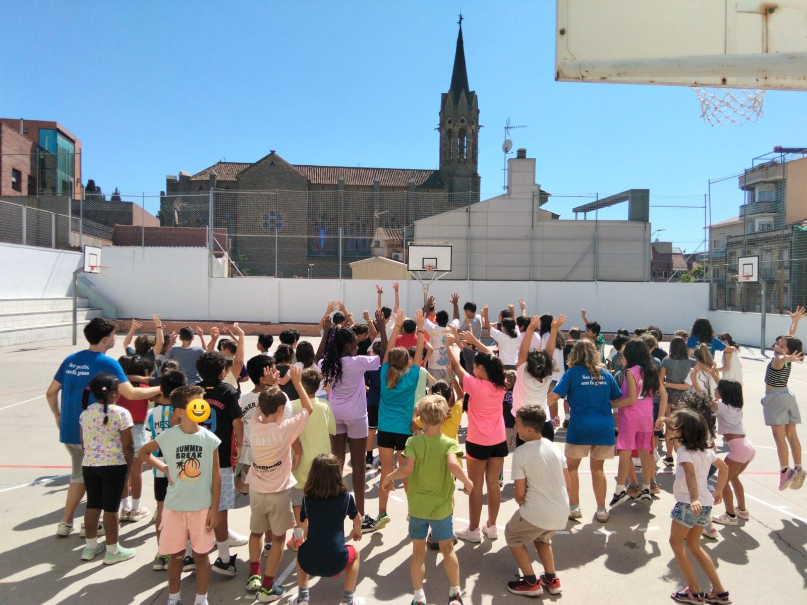 Un grup nombrós d’infants amb els braços enlaire formant una rotllana al pati, amb el campanar al fons.
