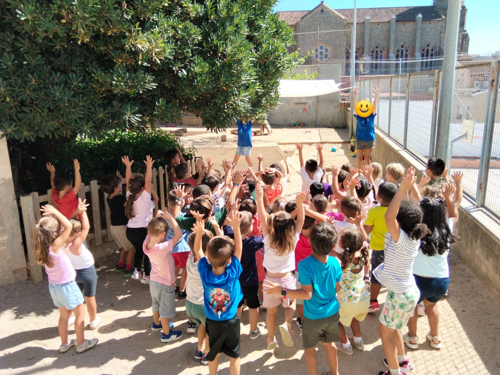 Infants amb els braços enlaire sota un arbre gran en una zona de terra del pati.