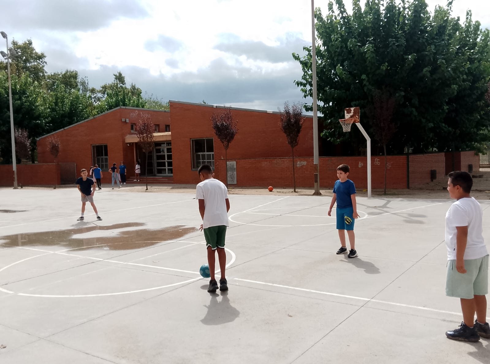 Infants jugant amb una pilota a la pista exterior, amb l’edifici de maó de l’escola al fons.
