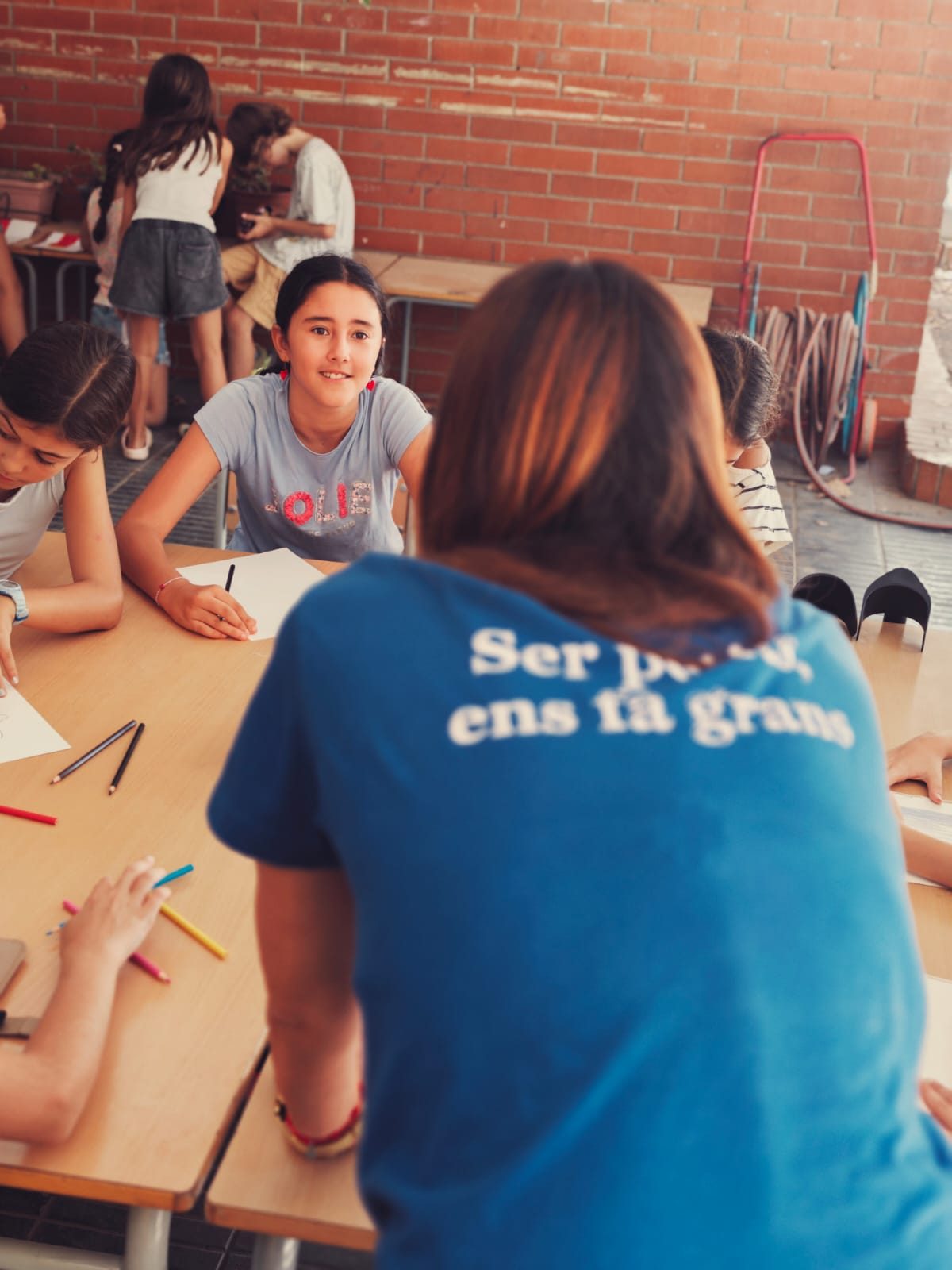Una educadora acompanya un grup d’infants que dibuixa en una taula del pati.