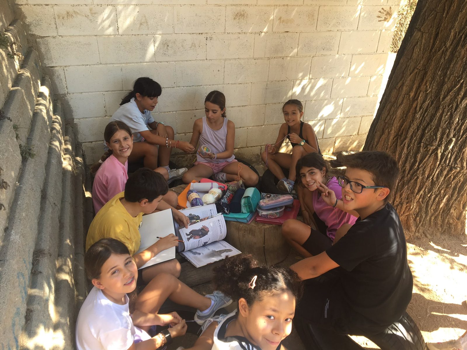 Un grup d’infants seu a terra a l’ombra, amb llibres i estoigs.