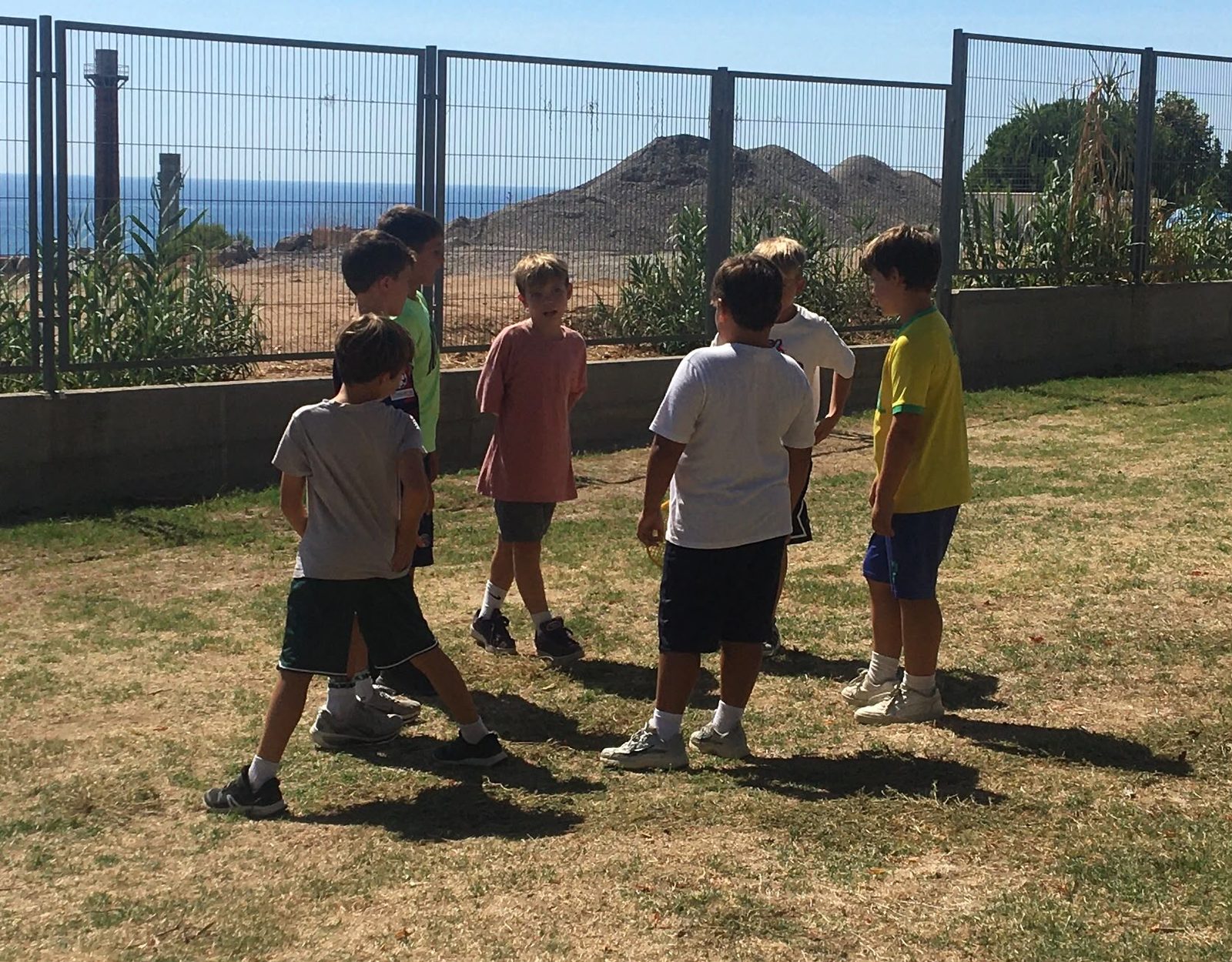 Infants jugant en una zona de gespa amb el mar al fons.