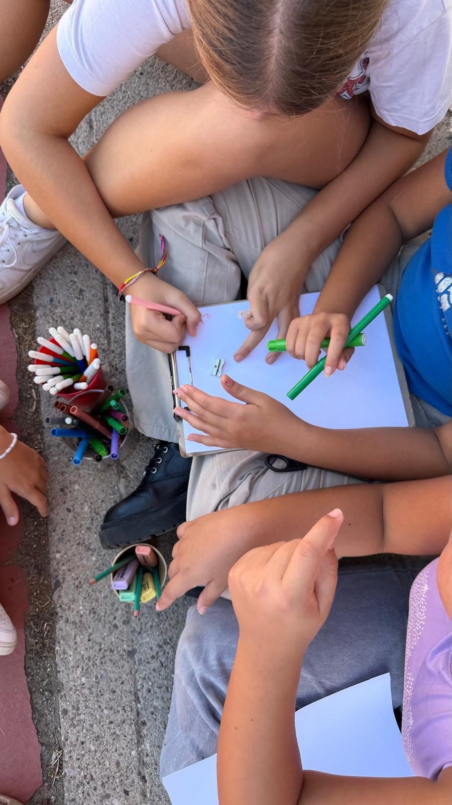 Infants dibuixen a terra amb un pot de retoladors al mig.