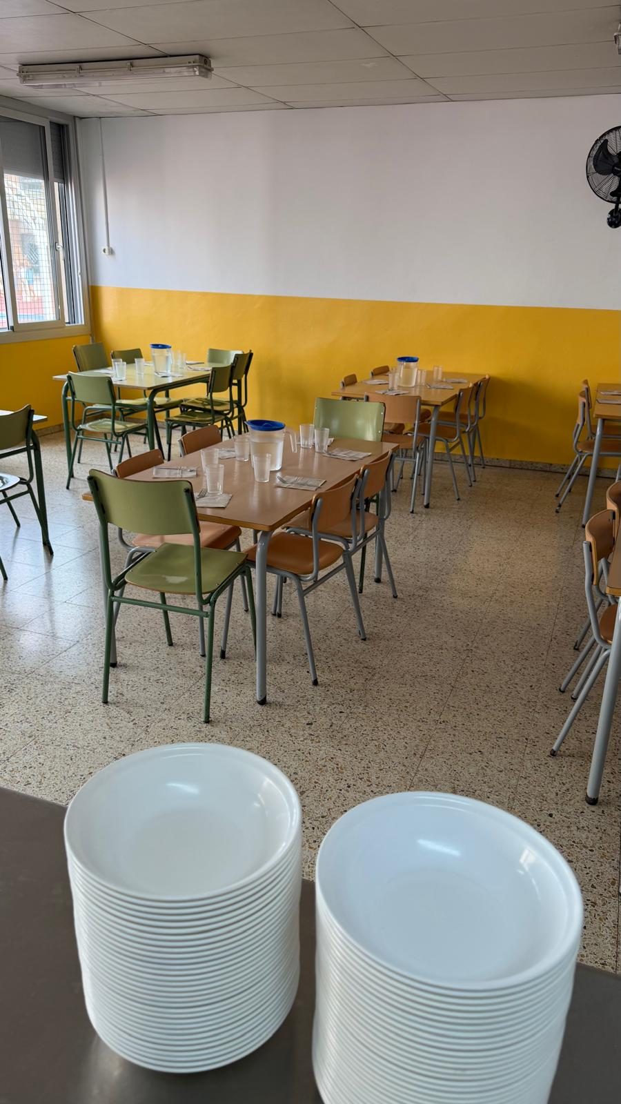 Menjador amb les taules parades amb gots i dues piles de plats blancs en primer terme.